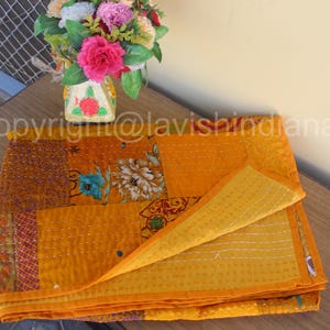 May include: A yellow kantha quilt with a floral pattern and a border of red and pink. The quilt is folded in half and is sitting on a wooden table. A vase of flowers is sitting on the table in the background.