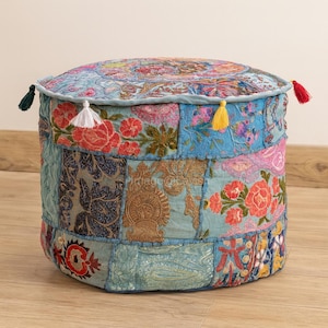 Puede incluir: Un puf redondo de patchwork con un diseño floral. El puf presenta una variedad de colores, incluyendo azul, rosa y dorado. Tiene un aspecto suave y texturizado y está adornado con borlas de varios colores.