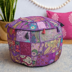 May include: A round, patchwork pouf in shades of purple, featuring a variety of floral and paisley patterns. The pouf has a flat top and bottom, with small tassels in various colors around the edges. The overall design is bohemian and decorative.
