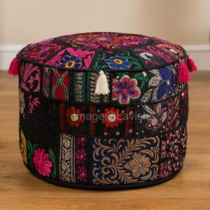 May include: A round, patchwork ottoman with a black base and a colourful, embroidered top. The design features floral and geometric patterns in shades of pink, blue, and gold. It has pink tassels and a white tassel.