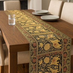 May include: A decorative table runner with an intricate pattern featuring elephants, birds, and floral motifs in gold, red, and black. The runner is placed on a dark wooden table, enhancing the rich colors and design. Plates and a glass of water are also on the table.