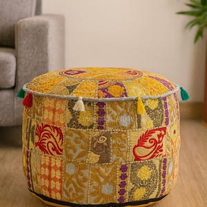 May include: Round, patchwork ottoman in shades of yellow, orange, red, and purple. The top features a circular design, and the sides are adorned with various geometric and floral patterns. Small tassels in multiple colors are attached around the top edge.