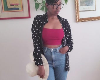May include: A woman wearing a black polka dot button-up shirt, a red crop top, and blue jeans. She is holding a white sun hat. The outfit is paired with black shoes. The background includes framed artwork and greenery.