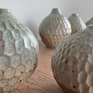 May include: A selection of handmade ceramic vases with a textured, honeycomb-like design. The vases are a blend of light green and beige, with a rounded form and a small opening at the top. They are displayed on a wooden surface.