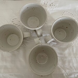 May include: Four speckled ceramic mugs with handles arranged in a circular pattern. The mugs are off-white with dark brown speckles, and sit on a white, textured surface. The mugs are empty and ready to be filled.