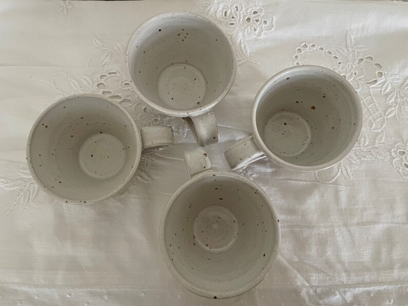 May include: Four speckled ceramic mugs with handles arranged in a circular pattern. The mugs are off-white with dark brown speckles, and sit on a white, textured surface. The mugs are empty and ready to be filled.