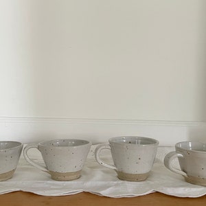 May include: Four speckled ceramic mugs with handles, each with a light beige base and a white upper portion. The mugs are arranged on a white, textured surface, likely a tablecloth, against a plain white wall.