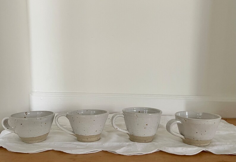 May include: Four speckled ceramic mugs with handles, each with a light beige base and a white upper portion. The mugs are arranged on a white, textured surface, likely a tablecloth, against a plain white wall.