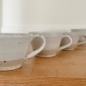 May include: Four handmade ceramic mugs with a speckled, off-white glaze and a natural, unglazed base. Each mug has a curved handle and a slightly flared rim. The mugs are arranged in a row on a wooden surface.