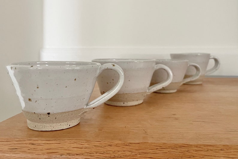 May include: Four handmade ceramic mugs with a speckled, off-white glaze and a natural, unglazed base. Each mug has a curved handle and a slightly flared rim. The mugs are arranged in a row on a wooden surface.