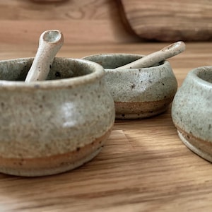 May include: A collection of four small, handmade ceramic bowls with accompanying spoons. The bowls are a speckled, muted green with a brown band at the base. The spoons share a similar colour and texture. The bowls are displayed on a wooden surface.