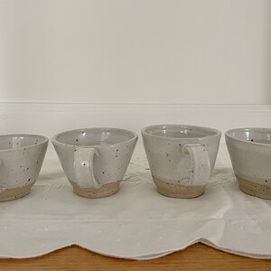 May include: Four speckled ceramic mugs with handles. The mugs are off-white with brown speckles and a natural, unglazed base. They are arranged in a row on a white, textured surface.