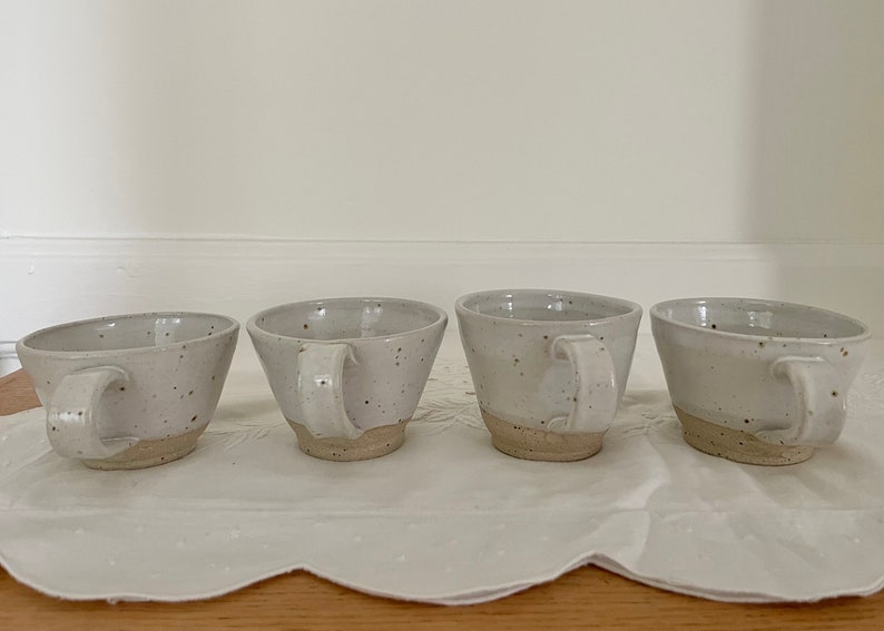 May include: Four speckled ceramic mugs with handles. The mugs are off-white with brown speckles and a natural, unglazed base. They are arranged in a row on a white, textured surface.