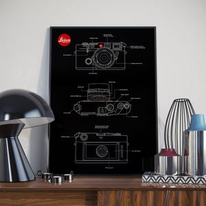 May include: A black and white line drawing of a Leica camera with labeled parts on a black background. The camera is shown in three different views: top, side, and bottom. The Leica logo is in red.