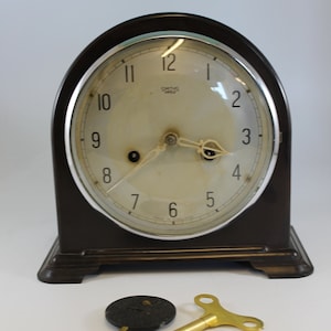Puede incluir: Un reloj Smiths vintage con una caja de madera negra y una esfera blanca. El reloj tiene un mecanismo de cuerda con llave y viene con una llave de cuerda.