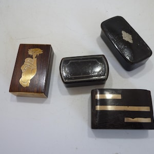 Könnte beinhalten: Vier rechteckige Schachteln in verschiedenen Größen und Materialien. Eine ist aus dunklem Holz mit einer goldenen Einlage einer Hand, die eine Blume hält. Eine weitere ist dunkelbraun mit drei horizontalen goldenen Metallstreifen. Zwei sind schwarz mit dekorativen Metallakzenten.