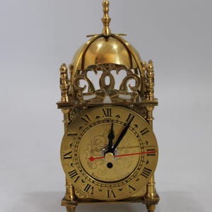 Può includere: Un orologio in ottone dorato con una cima a forma di cupola e un quadrante con numeri romani. L'orologio è in stile tradizionale inglese a pendolo.