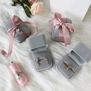 Peut inclure: Quatre bo&icirc;tes &agrave; bijoux en velours gris avec des accents de ruban rose. Deux bo&icirc;tes sont ouvertes et pr&eacute;sentent des boucles d'oreilles et une bague. Une bo&icirc;te est ferm&eacute;e et a un n&oelig;ud de ruban rose. La quatri&egrave;me bo&icirc;te est ferm&eacute;e et a un n&oelig;ud de ruban rose avec un charme de perle.