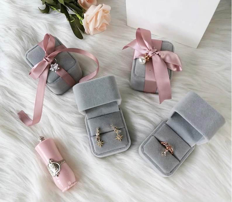 Peut inclure: Quatre bo&icirc;tes &agrave; bijoux en velours gris avec des accents de ruban rose. Deux bo&icirc;tes sont ouvertes et pr&eacute;sentent des boucles d'oreilles et une bague. Une bo&icirc;te est ferm&eacute;e et a un n&oelig;ud de ruban rose. La quatri&egrave;me bo&icirc;te est ferm&eacute;e et a un n&oelig;ud de ruban rose avec un charme de perle.