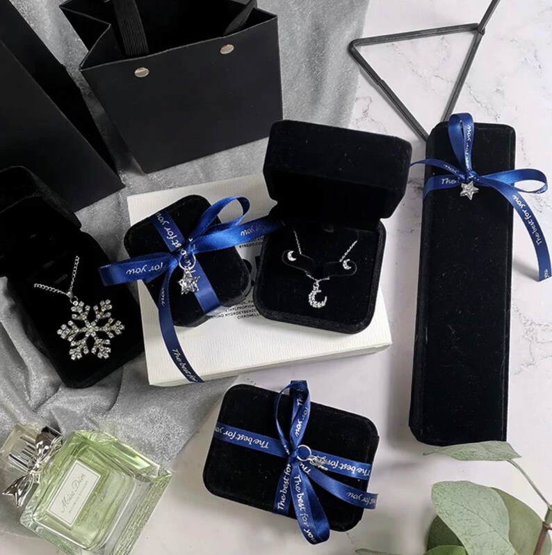 Peut inclure: Cinq bo&icirc;tes &agrave; bijoux en velours noir avec des n&oelig;uds en ruban bleu. Les bo&icirc;tes sont sur une surface blanche avec un flacon de parfum et des feuilles vertes. Le texte "The best for you" est imprim&eacute; sur les bo&icirc;tes.