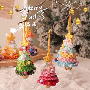 Puede incluir: Cuatro adornos de árbol de Navidad de crochet de colores vivos con pompones y una campana en cada uno. Los adornos están colgados de una rama con un copo de nieve blanco y las palabras "Merry Christmas" escritas en blanco.