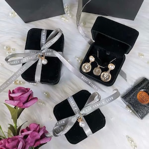 Peut inclure: Trois bo&icirc;tes cadeaux en velours noir avec un ruban argent&eacute; nou&eacute; en n&oelig;uds. Le ruban porte l'inscription "Best Wishes For You". Les bo&icirc;tes sont pos&eacute;es sur une surface blanche et moelleuse avec des perles blanches &eacute;parpill&eacute;es autour. Une bo&icirc;te contient un collier et des boucles d'oreilles en or avec des accents de perles.