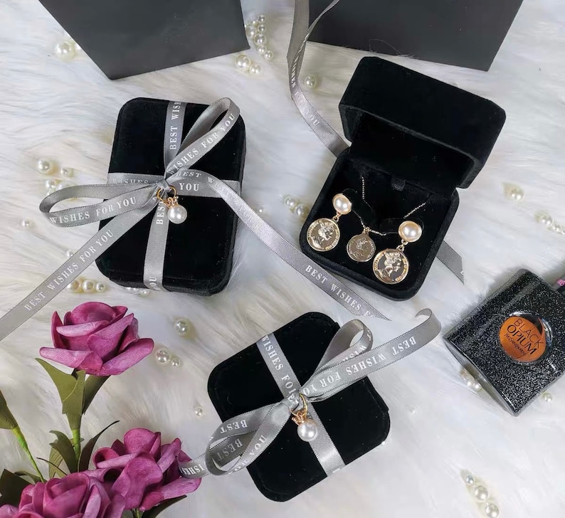Peut inclure: Trois bo&icirc;tes cadeaux en velours noir avec un ruban argent&eacute; nou&eacute; en n&oelig;uds. Le ruban porte l'inscription "Best Wishes For You". Les bo&icirc;tes sont pos&eacute;es sur une surface blanche et moelleuse avec des perles blanches &eacute;parpill&eacute;es autour. Une bo&icirc;te contient un collier et des boucles d'oreilles en or avec des accents de perles.