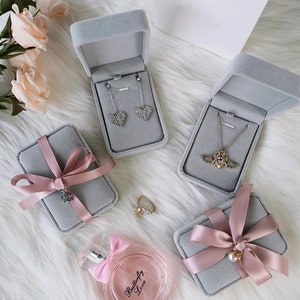 Peut inclure: Quatre bo&icirc;tes &agrave; bijoux en velours gris avec des n&oelig;uds en ruban rose. Deux bo&icirc;tes contiennent des colliers, une contient des boucles d'oreilles et une contient une bague. Les bo&icirc;tes sont sur une surface blanche et moelleuse.