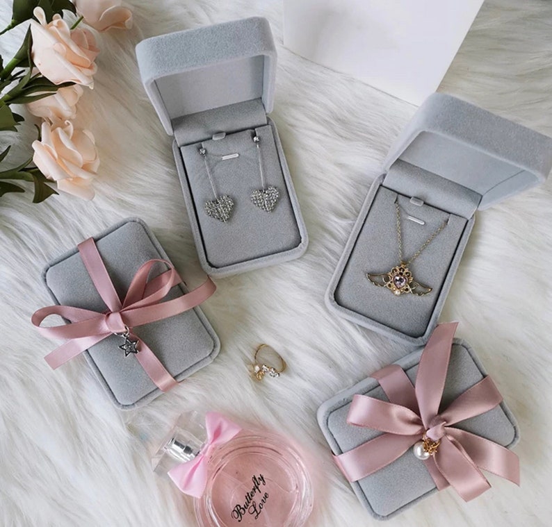 Peut inclure: Quatre bo&icirc;tes &agrave; bijoux en velours gris avec des n&oelig;uds en ruban rose. Deux bo&icirc;tes contiennent des colliers, une contient des boucles d'oreilles et une contient une bague. Les bo&icirc;tes sont sur une surface blanche et moelleuse.
