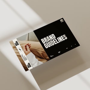 May include: A black and white brand guideline document with the text "BRAND GUIDELINES" in bold white letters. The document is lying on a light brown surface with shadows from a window.
