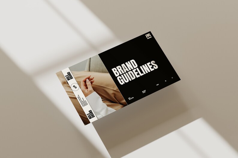 May include: A black and white brand guideline document with the text "BRAND GUIDELINES" in bold white letters. The document is lying on a light brown surface with shadows from a window.