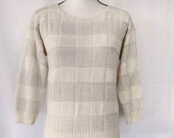 May include: A cream-colored sweater with a checkered pattern. The sweater has a round neckline and long sleeves.