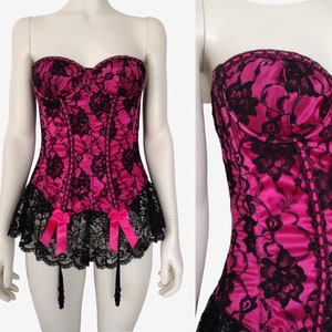 May include: A pink satin and black lace corset with a sweetheart neckline and a black lace trim. The corset has a boned structure and a matching garter belt with pink bows.