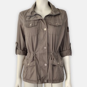 May include: A taupe-colored jacket with a stand-up collar, zipper, and button closures. The jacket features four front pockets, adjustable waist drawstring, and rolled-up sleeves. An American flag patch is visible on the upper left sleeve.