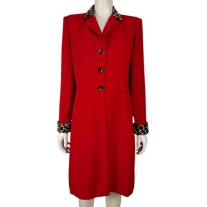 May include: A long, bright red coat featuring a structured collar and cuffs with a brown and cream giraffe-inspired animal print pattern. The coat has three black buttons down the center front, long sleeves, and a straight, knee-length hemline.