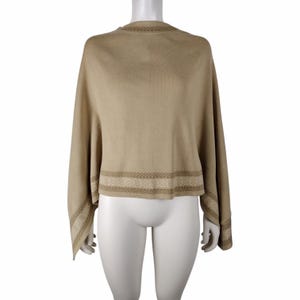 May include: A tan poncho-style sweater with long sleeves. The sweater has a decorative geometric pattern in a lighter shade of tan along the bottom hem and sleeve cuffs. The sweater is displayed on a white mannequin.