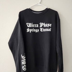 Peut inclure: Sweat-shirt noir avec un texte blanc qui dit "Wicca Phase Springs Eternal" sur le devant et "Phase" sur la manche gauche.
