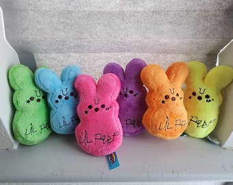 Könnte beinhalten: Sechs bunte Plüsch-Osterhasen mit dem Text "Lil Peep" auf jedem. Die Hasen sind grün, blau, rosa, lila, orange und gelb.