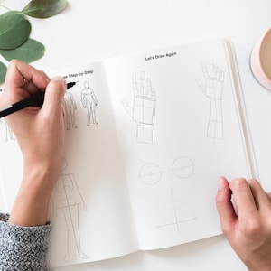 Pode incluir: Caderno de esboços aberto com tutoriais de desenho de figuras e mãos. Uma mão segurando uma caneta preta desenha uma figura. Os textos "Artline" e "Let's Draw Again" são visíveis. Uma caneca rosa com uma bebida clara e folhas de eucalipto também estão na imagem.