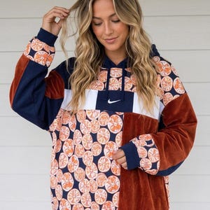May include: A long-sleeve hooded sweatshirt featuring a patchwork design of navy blue fabric, brown corduroy panels, and a repeating orange citrus slice print. The garment includes a white horizontal stripe and a small white Nike logo on the chest.