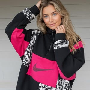 May include: Black, pink, and floral patterned Nike sweatshirt with a quarter-zip collar. The sweatshirt features a large pink panel across the chest with the Nike swoosh logo. The sleeves and cuffs have a black and white floral design.