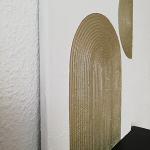 Peut inclure: Une toile blanche avec deux formes abstraites peintes en or. Les formes sont une grande arche et un demi-cercle plus petit.