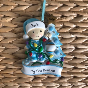 May include: Blue and white Christmas ornament featuring a cartoon baby boy wearing a blue and white pajamas with a string of lights, holding a blue and white ornament. The ornament has a banner that reads "My First Christmas" and the baby's name "Bart" is written on his hat. The year "2020" is written on the top of the Christmas tree.