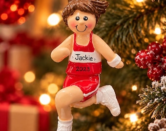 May include: A hanging ornament of a runner with brown hair in a ponytail, wearing a red and white tank top and shorts with white athletic shoes. The shirt features the name Jackie and the year 2023 printed in black text. A red ribbon is attached.