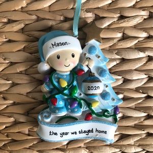 May include: A blue and white Christmas ornament featuring a baby boy in pajamas holding a blue ornament with the year 2020 on it. The ornament is decorated with a blue Christmas tree and lights. The text "the year we stayed home" is on the bottom of the ornament.
