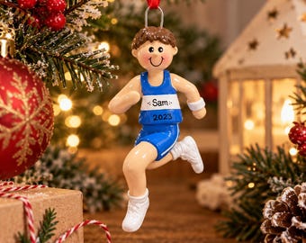 May include: A hanging ornament of a runner with brown hair, wearing a blue and white tank top with the name Sam and the year 2023 printed on the front, blue shorts, and white shoes, suspended by a red ribbon from a metal loop.