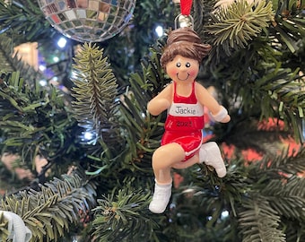 Puede incluir: Adorno navideño que representa a una corredora con uniforme rojo con el nombre "Jackie" y el año "2023". El adorno cuelga de un árbol de Navidad verde con otros adornos, incluyendo una bola de discoteca plateada.