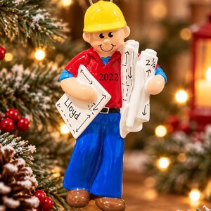 May include: A hanging ornament of a construction worker wearing a yellow hard hat, red shirt, and blue trousers. The figure holds white blueprints with the name Lloyd and the year 2022 printed on them. The ornament features a red ribbon loop.