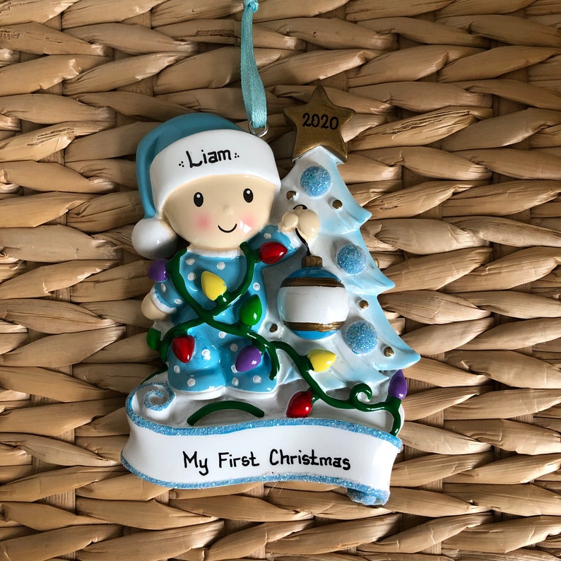 May include: A blue and white Christmas ornament featuring a baby in pajamas holding a Christmas tree ornament. The ornament has the text "Liam" and "My First Christmas" on it. The year "2020" is on the top of the tree.
