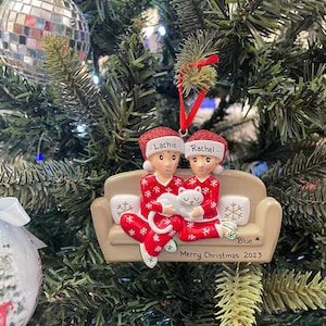 Op de afbeelding: Een kerstboomornament met twee figuren in rode pyjama's met sneeuwvlokpatronen, zittend op een beige bank en een witte kat vasthoudend. Het ornament heeft de namen "Lachie" en "Rachel" en de tekst "Merry Christmas 2023".